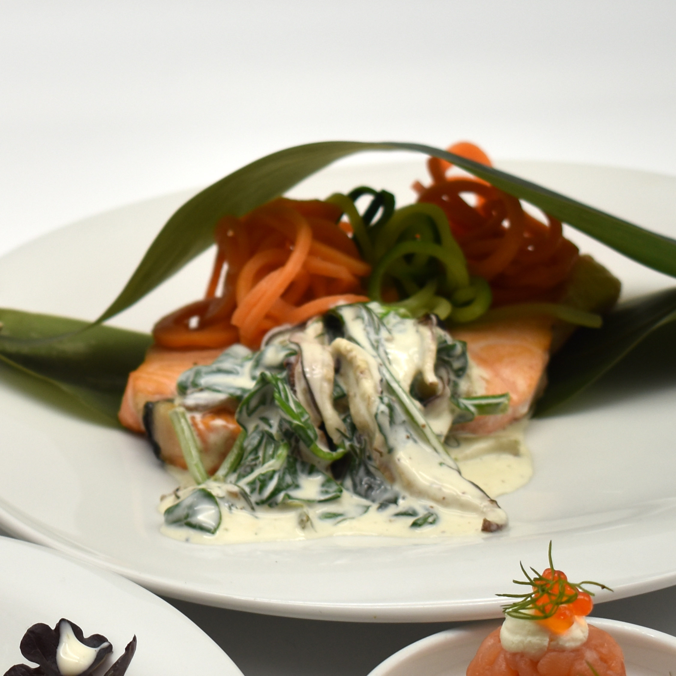 Medallón de salmón especiado en Papillote de hoja de Coco con tallarines de zanahoria, calabacín, batata y una crema de espinacas y champiñones shiitake.